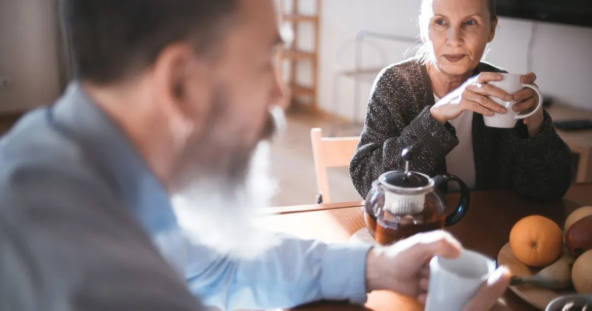 Older man and woman sharing tea at a wooden table with a bowl of fruit.