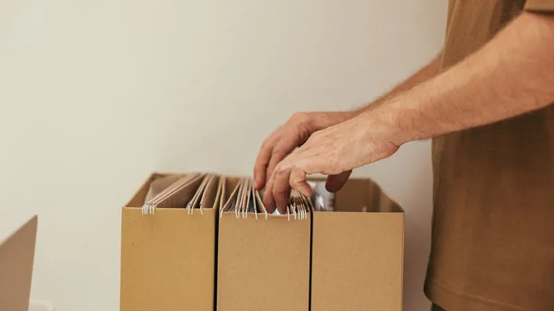 A person's hands flipping through hanging file folders in cardboard boxes.