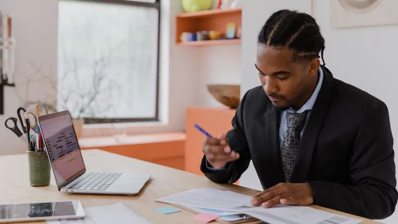 A man in a suit and tie reviews paperwork at a desk beside a laptop.