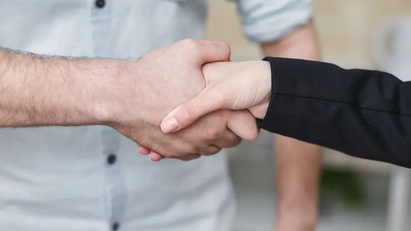 Close-up of a man in a light shirt shaking hands with a woman in a black blazer.