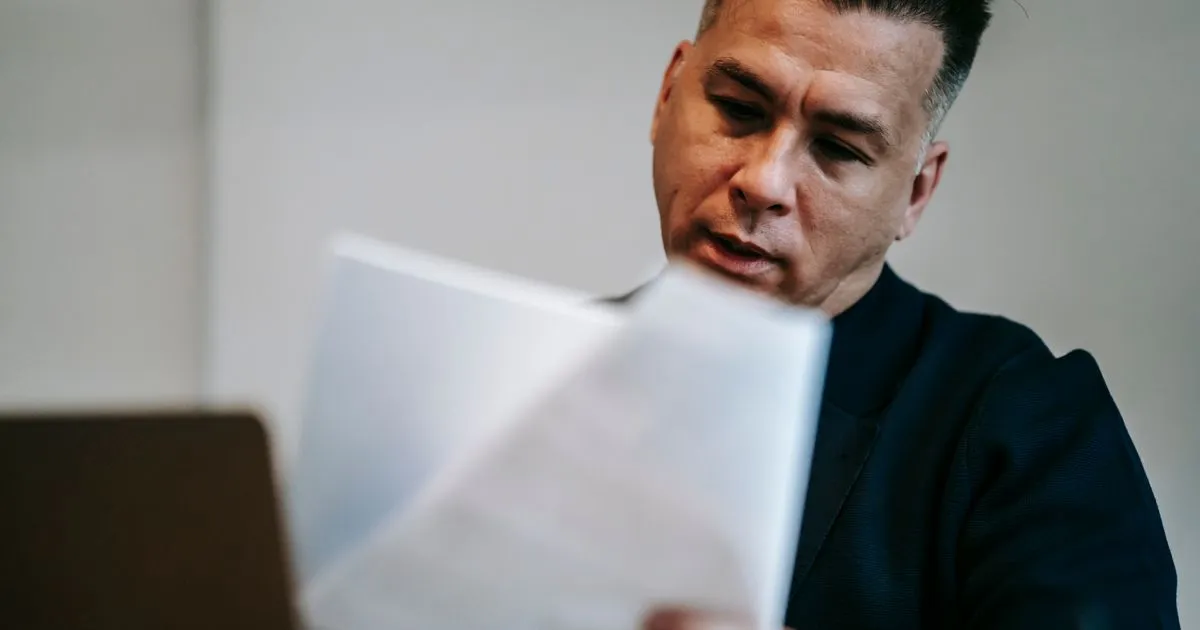 Man in a dark blazer reads printed documents beside an open laptop.