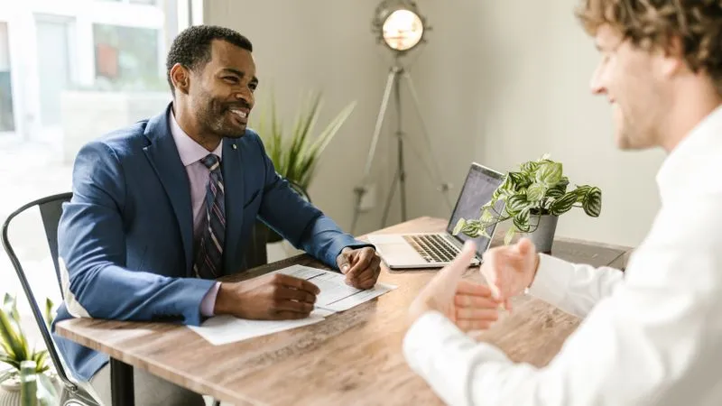 Two men talk across a wooden table with papers, a laptop and a potted plant.