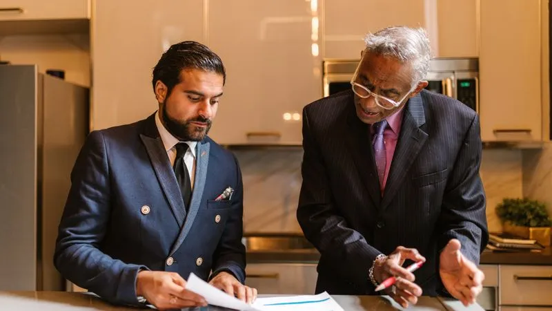 Two men in suits review paperwork together at a kitchen counter, one holding a pen.