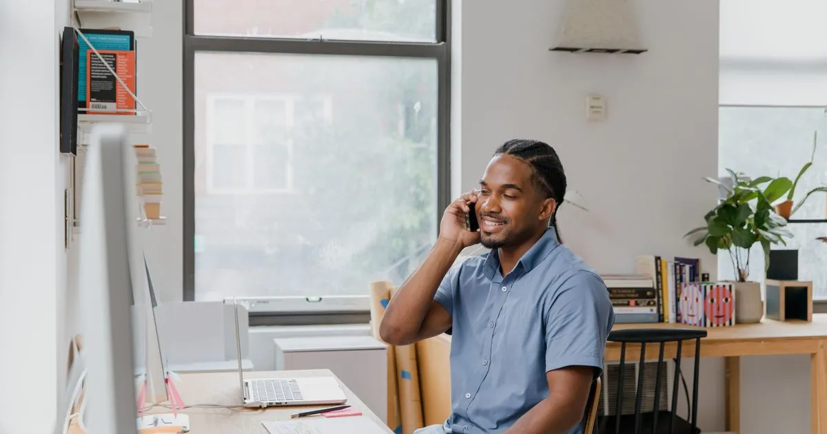Man in a blue shirt smiles while talking on the phone at a desk with a laptop.