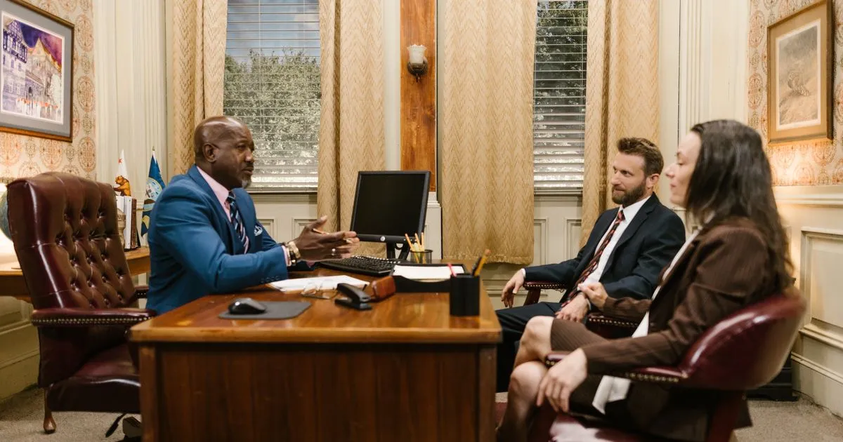A man in a blue suit at a desk talks with a man and woman seated opposite.