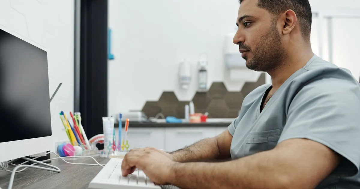A dentist in gray scrubs types on a computer keyboard at a clinic desk.