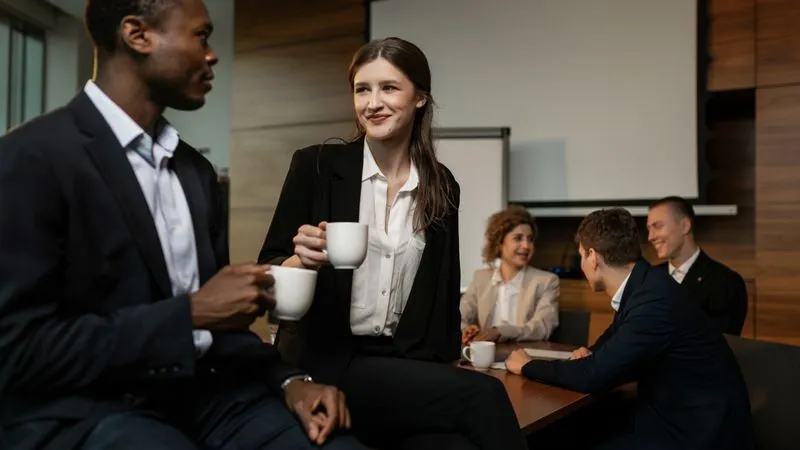 Colleagues in business attire chat over coffee cups in a wood-paneled conference room.
