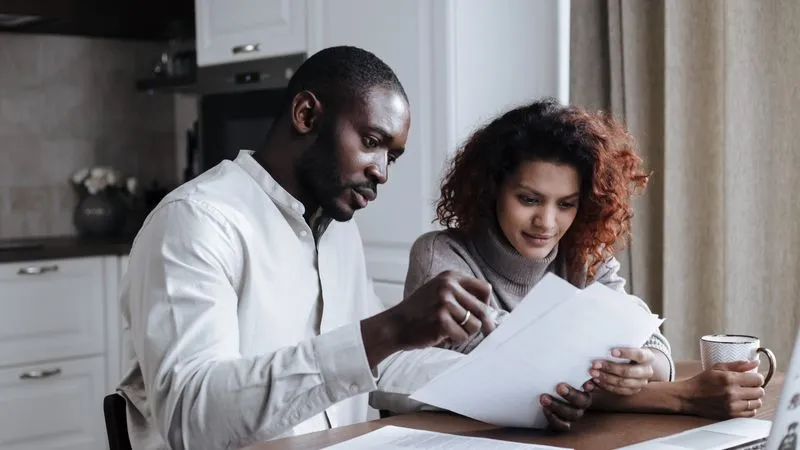 A man and woman review paperwork together at a kitchen table with a laptop.
