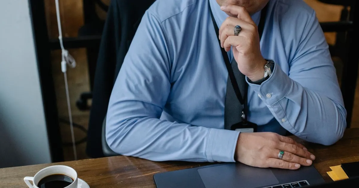 Man in a blue shirt and tie sits thoughtfully at a desk with a laptop and coffee.
