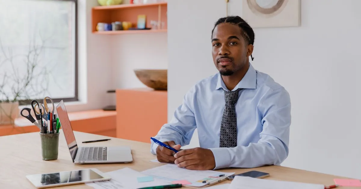 A man in a shirt and tie sits at a desk with a laptop and papers.