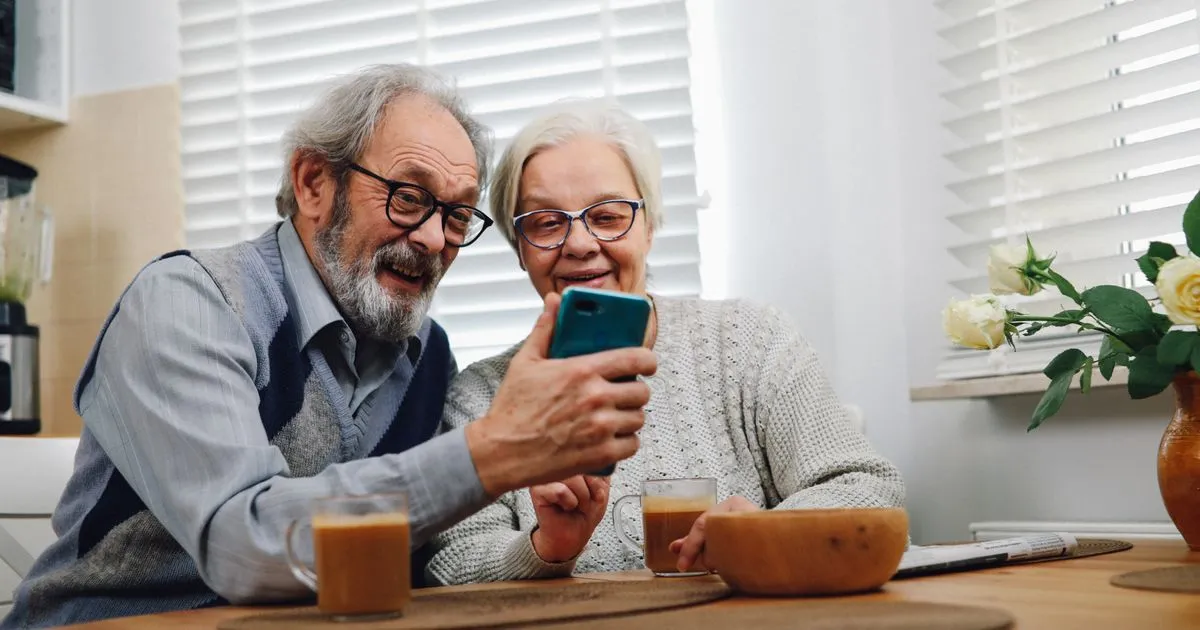 Older couple at a kitchen table with coffee, smiling at a smartphone together.